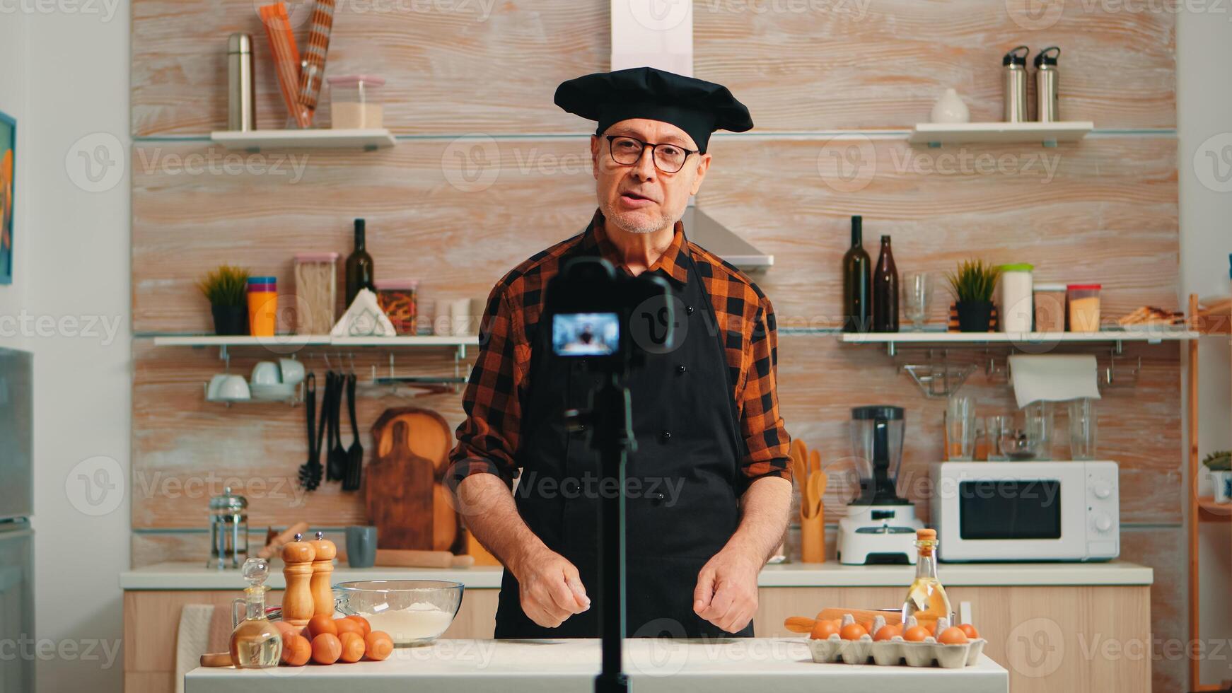 Elderly baker recording video tutorial about food recipe in kitchen. Retired blogger chefinfluencer using internet technology communicating, shooting, blogging on social media with digital equipment photo