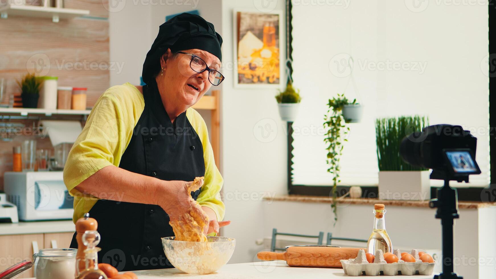 Mature experienced baker recording culinary recipe tutorial using modern camera in kitchen wearing bonete and apron. Influencer chef using internet technology communicating on social media. photo