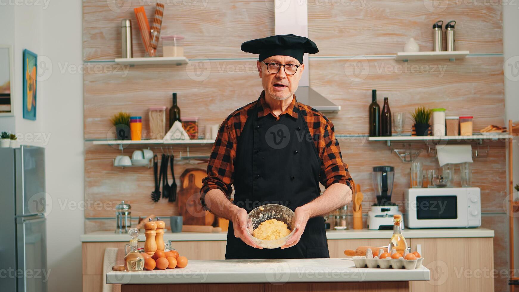 Old man wearing bonete while explaining food preparation recipe looking at camera. Retired blogger chef influencer using internet technology communicating on social media with digital equipment photo