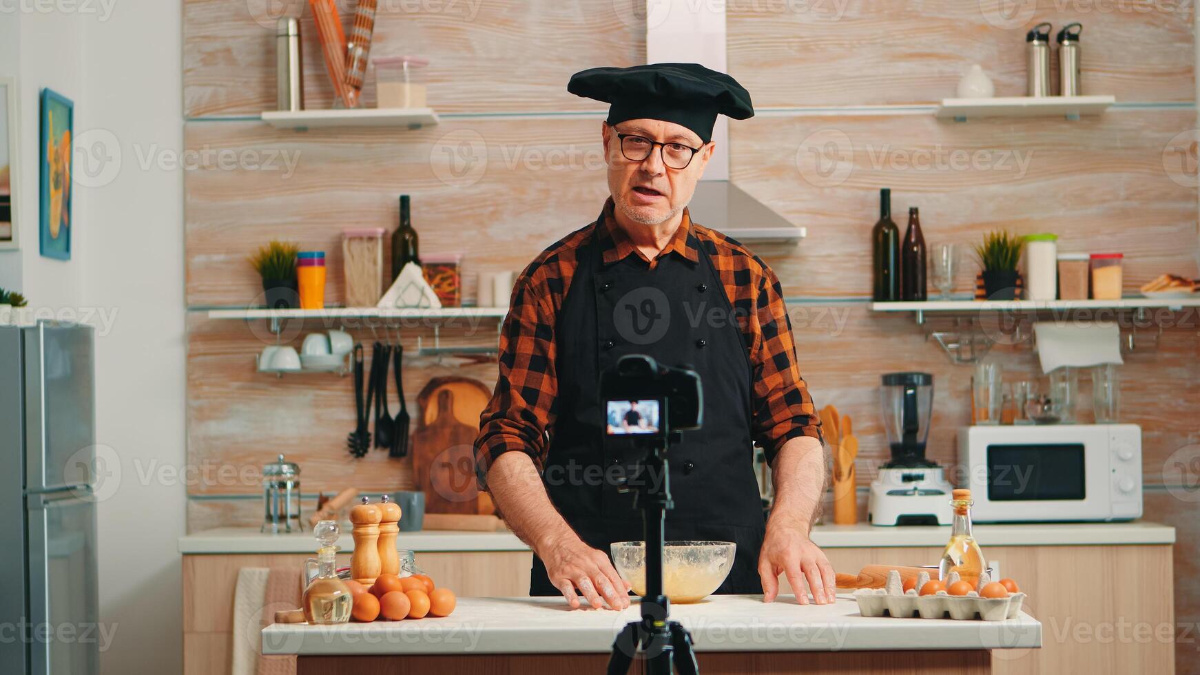 Mature experienced baker explaining recipe for audience recording tutorial for social media. Retired blogger chef influencer using internet technology communicating, shooting with digital equipment photo