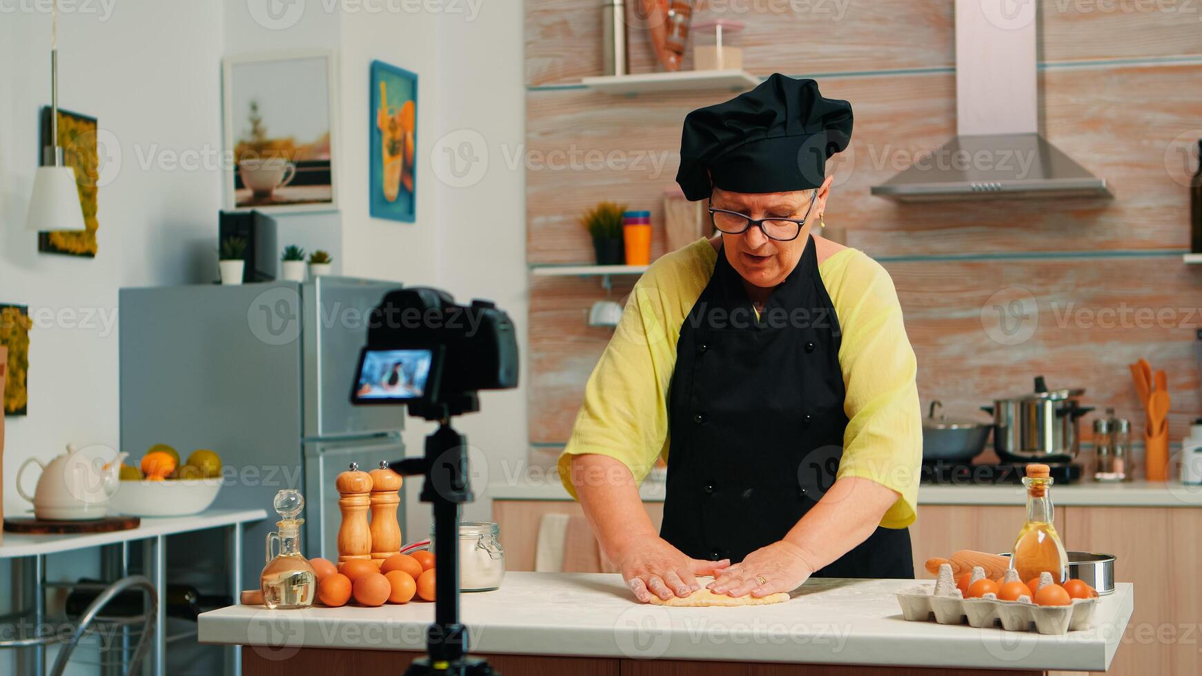 Old baker recording culinary video in kitchen preparing homemade pizza. Retired blogger chef influencer using internet technology communicating shooting blogging on social media with digital equipment photo