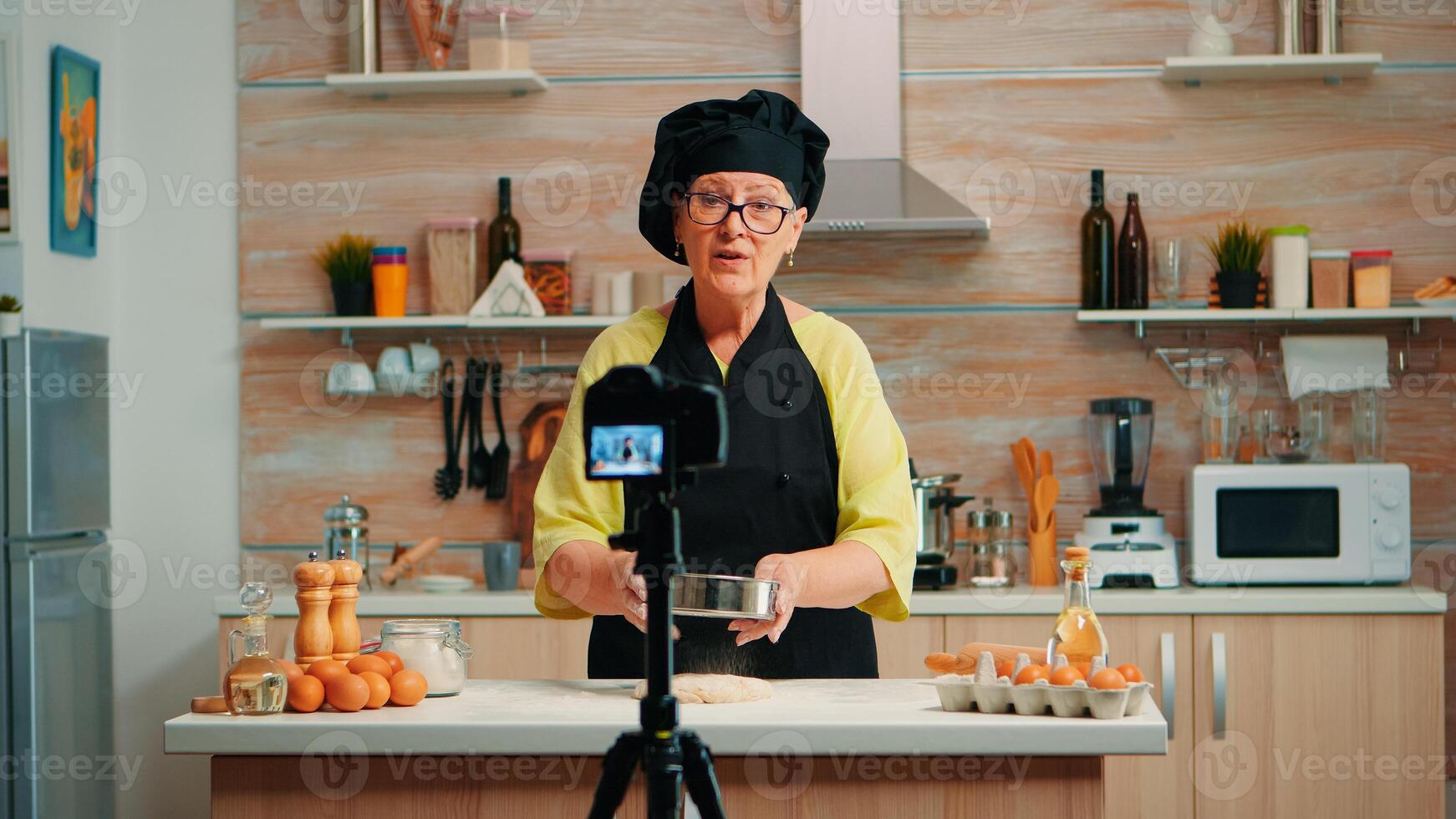 Happy elderly baker woman filming cooking vlog in home kitchen. Retired blogger chef influencer using internet technology communicating, shooting blogging on social media with digital equipment photo