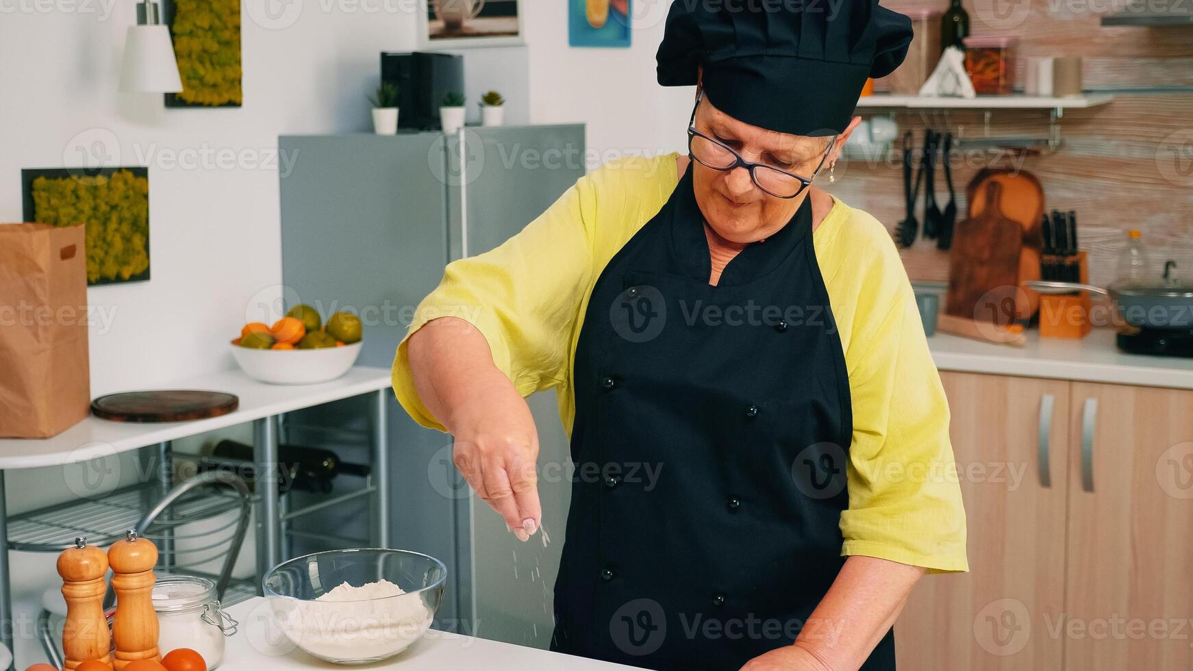 Woman chef taking wheat flour from glass bowl and sieving on table. Retired senior baker with bonete and uniform sprinkling, sifting, spreading rew ingredients baking homemade pizza and bread. photo