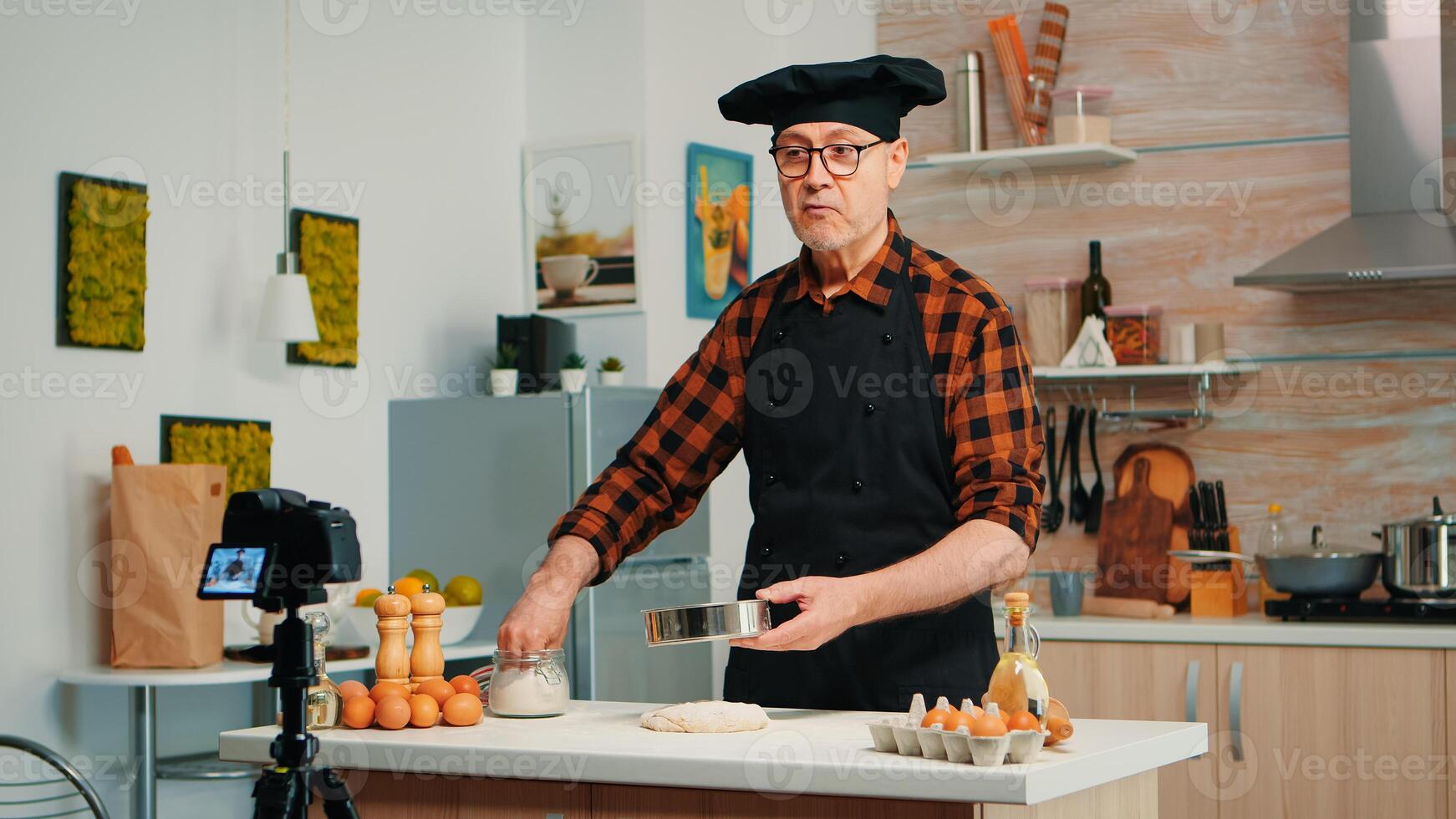 Cheerful elderly man recording the recipe step by step in hoe kitchen. Retired blogger chef influencer using internet technology communicating, shooting blogging on social media with digital equipment photo