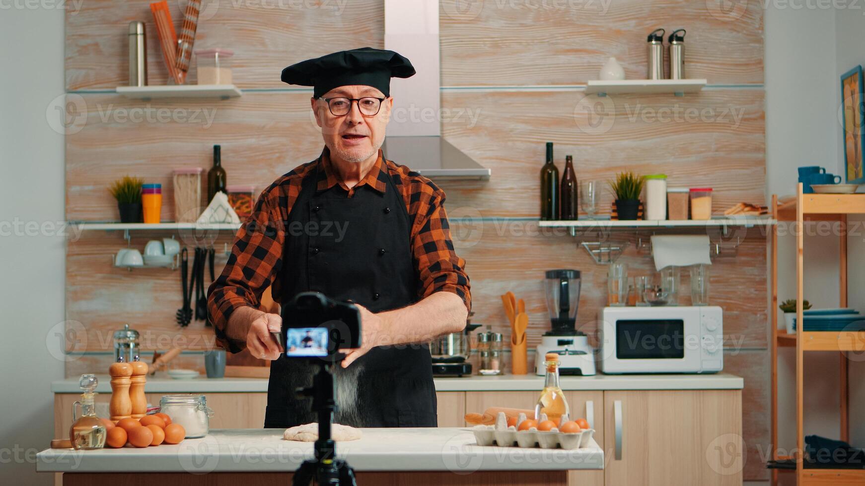 Cheerful elderly baker man filming cooking vlog in home kitchen. Retired blogger chef influencer using internet technology communicating, shooting blogging on social media with digital equipment photo