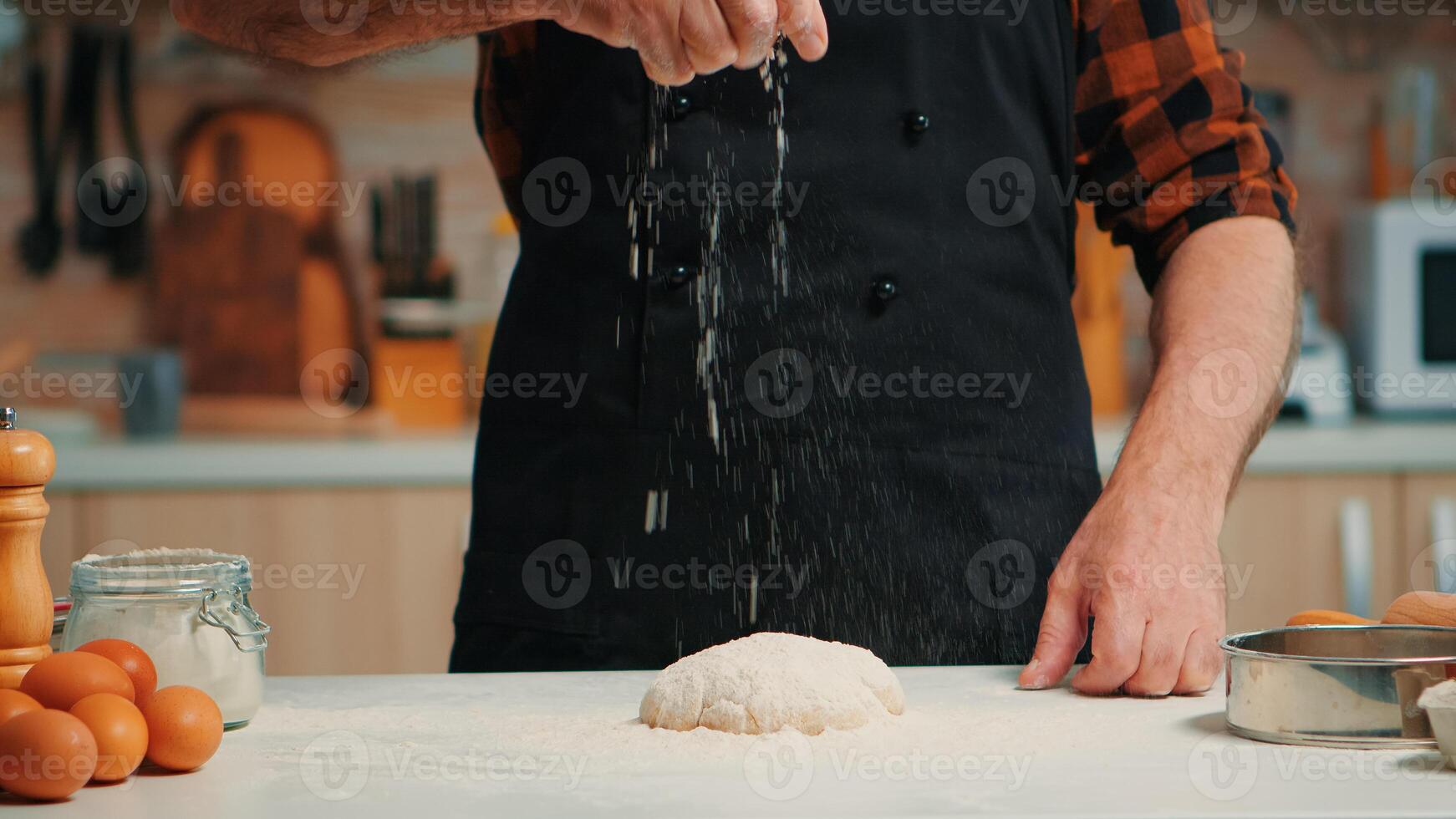 Senior bakery man sifting wheat flour on dough for kneading. Retired elderly chef with bonete and uniform sprinkling, sieving spreading rew ingredients with hand baking homemade pizza and bread. photo