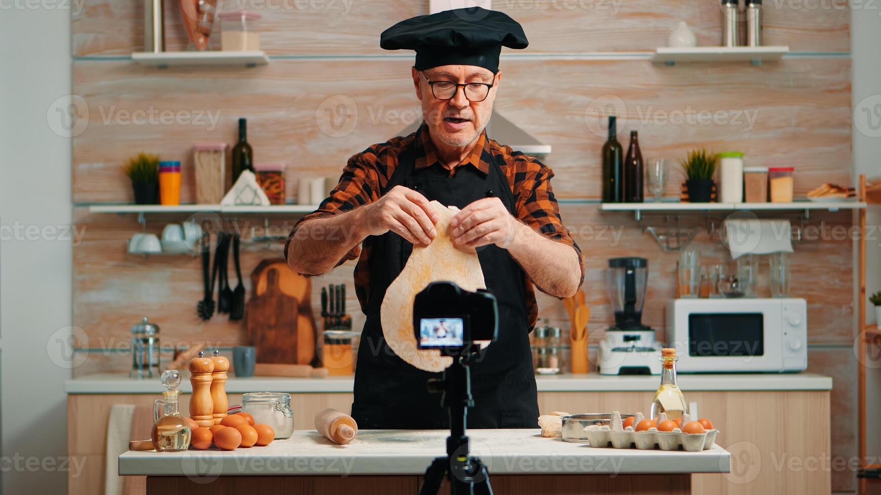 Baker using wooden rolling pin for dough in front video camera recording new cooking episode. Old blogger chef influencer using internet technology communicating on social media with digital equipment photo