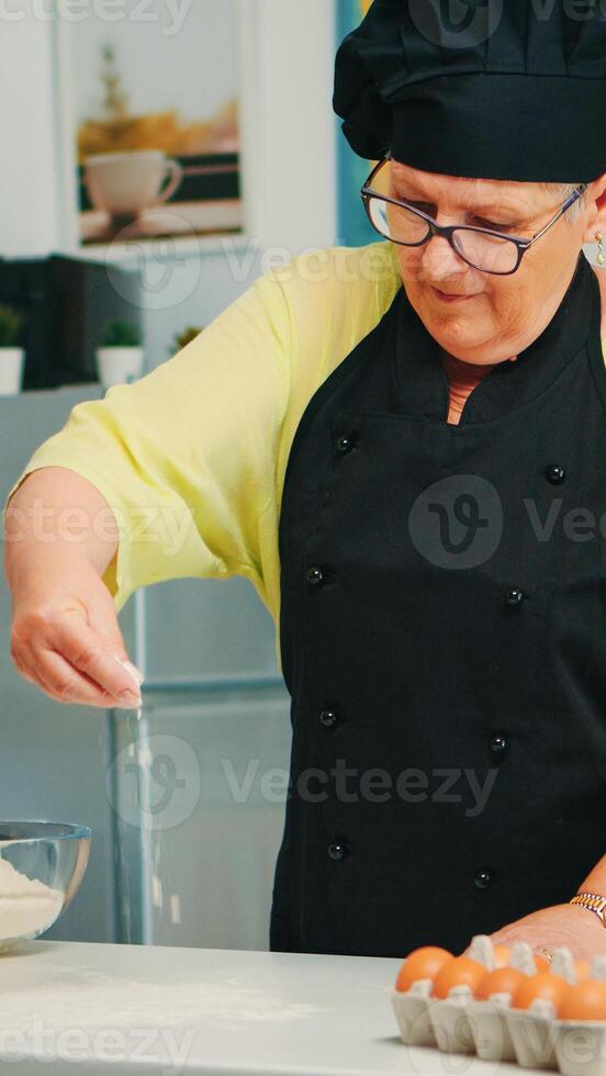 Baker spreading flour on wooden table at home in modern kitchen wearing apron and bonete. Happy elderly chef with uniform sprinkling, sieving sifting raw ingredients by hand baking homemade pizza photo