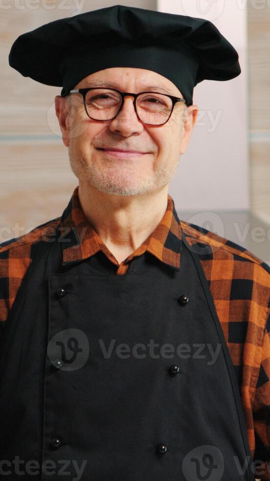 Senior baker smiling looking at camera in kitchen with food ingredients on table for baking. Retired elderly chef with bonete ready to cook homemade bread, cakes and pasta with wheat flour and eggs photo