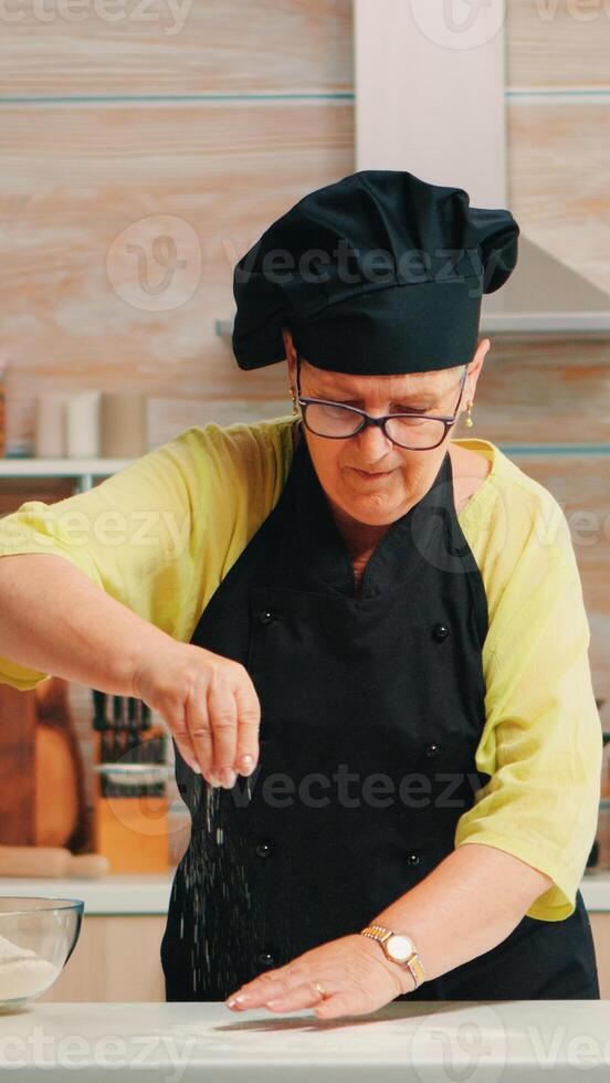 Woman is sifting flour on wooden table in modern kitchen. Happy elderly baker with bonete preparing raw ingredients to baking homemade cake sprinkling, sieving wheat flour by hand photo