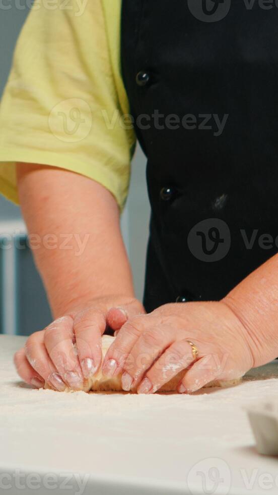 Woman hands kneading dough on table, preparing pastry and kitchen products. Retired elderly baker with bonete mixing ingredients with wheat flour for baking homemade cake and bread photo