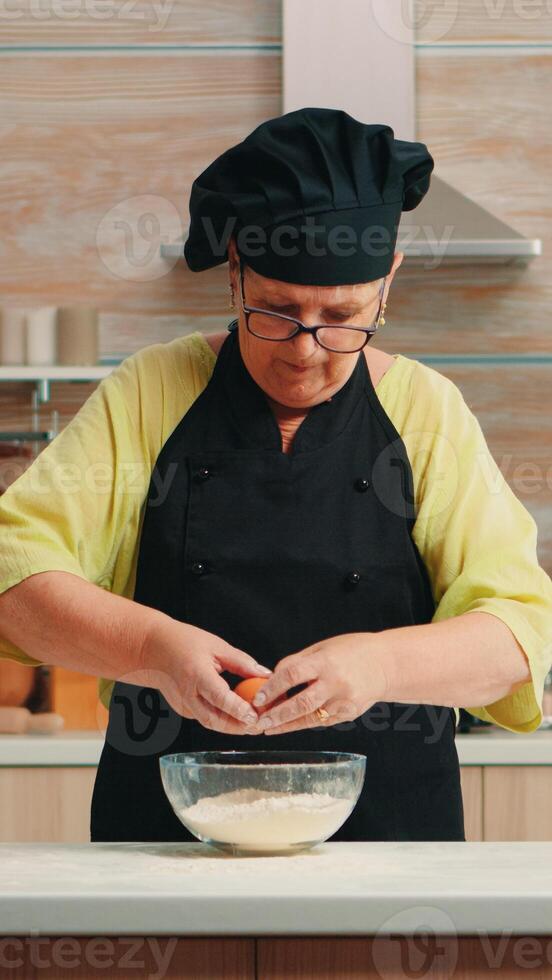 Woman baker cracking eggs into flour following traditional recipe in home kitchen. Retired elderly chef with bonete, mixing by hand, kneading in glass bowl pastry ingredients baking homemade cake photo