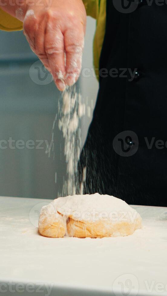 Grandmother preparing homemade donuts wearing kitchen apron. Retired senior chef with bonete and uniform sprinkling, sieving sifting wheat flour with hand baking homemade pizza and bread. photo