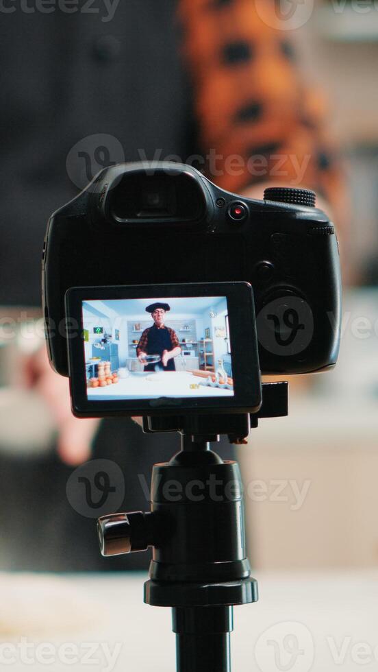 Close up of a video camera filming senior smiling man blogger in kitchen cooking. Retired blogger chef influencer using internet technology communicating on social media with digital equipment photo