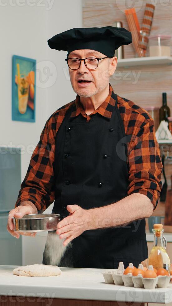 Cheerful elderly man recording the recipe step by step in hoe kitchen. Retired blogger chef influencer using internet technology communicating, shooting blogging on social media with digital equipment photo