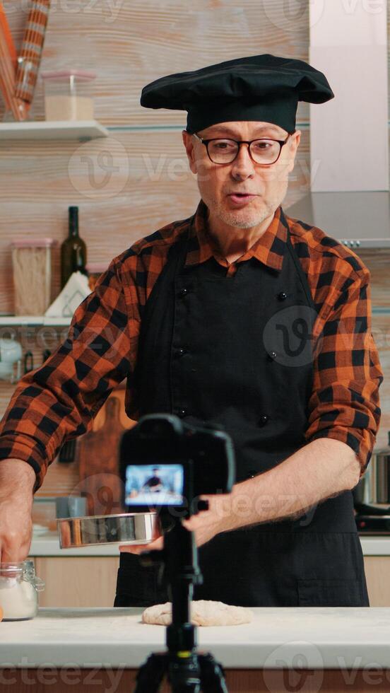 Cheerful elderly baker man filming cooking vlog in home kitchen. Retired blogger chef influencer using internet technology communicating, shooting blogging on social media with digital equipment photo