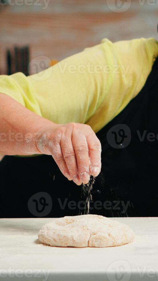 mujer con bonete y cocina delantal es ocupado con masa preparación. retirado mayor panadero con delantal, cocina uniforme aspersión, cernido, extensión harina con mano horneando hecho en casa Pizza y un pan. foto