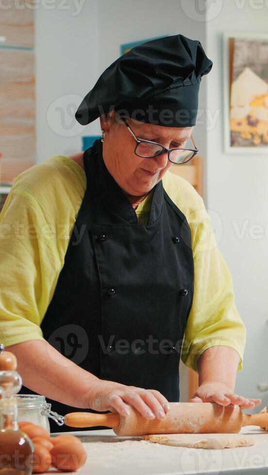 retirado mujer cocinero utilizando de madera laminación alfiler para Cocinando Pizza. contento mayor panadero con bonete preparando crudo ingredientes a horneando tradicional Pizza aspersión, tamizado harina en mesa en el cocina. foto