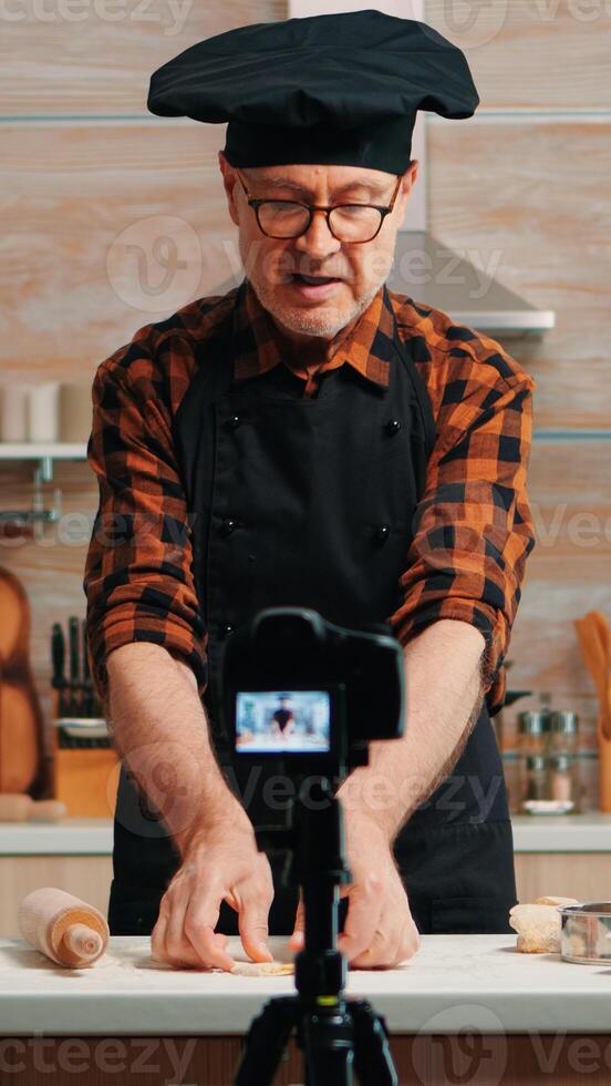 Baker using wooden rolling pin for dough in front video camera recording new cooking episode. Old blogger chef influencer using internet technology communicating on social media with digital equipment photo