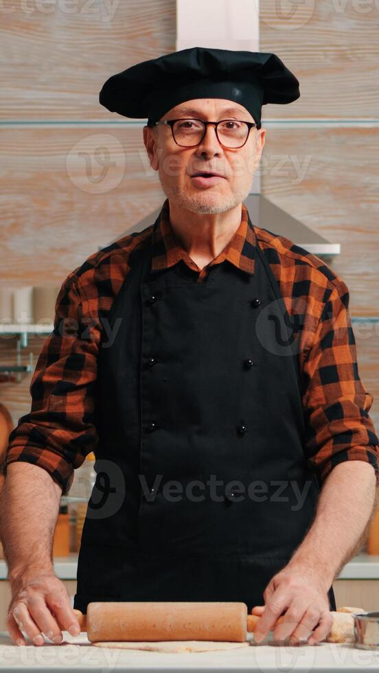 Elderly man rolling dough at home infront of video camera explaining recipe step by step. Retired blogger chef influencer using internet technology communicating on social media with digital equipment photo
