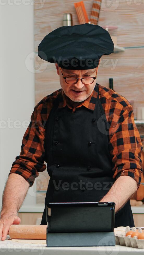 Retired man following culinary advice on tablet, learning cooking tutorial on social media, forming dough with wooden rolling pin. Grandfather with bonete and apron using laptop preparing home cakes photo