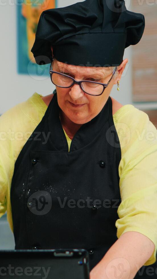 Cheerful retired chef in uniform spinning and tossing pizza dough throwing it up, forming on a floured surface and kneading it shaping. Skillful elderly baker wearing bonete cooking homemade cookies photo