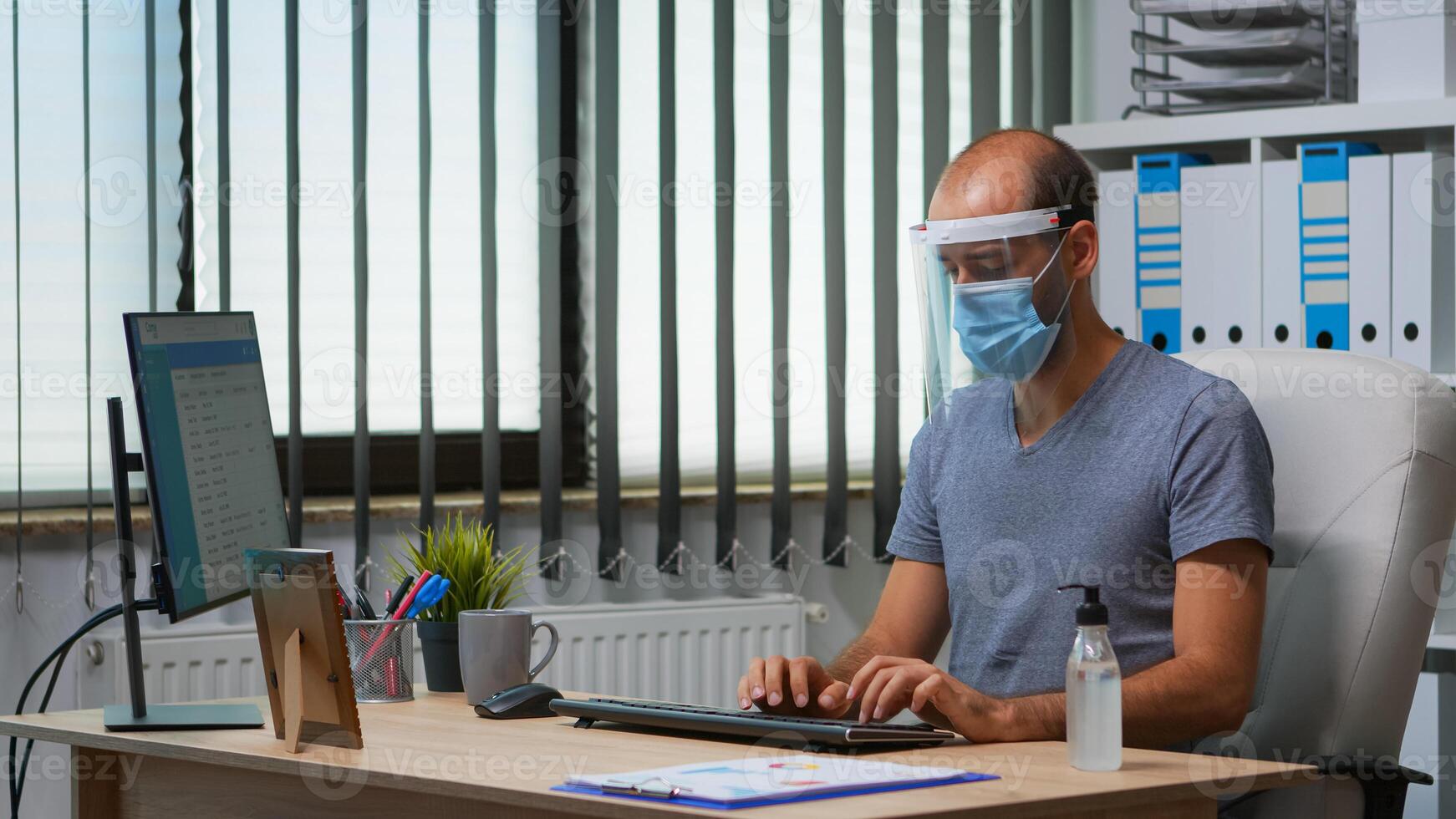 Man with protection mask and visor applying sanitizer gel rubbing hands before working at computer. Entrepreneur in new normal workplace disinfecting using antibacterial alcohol against corona virus. photo