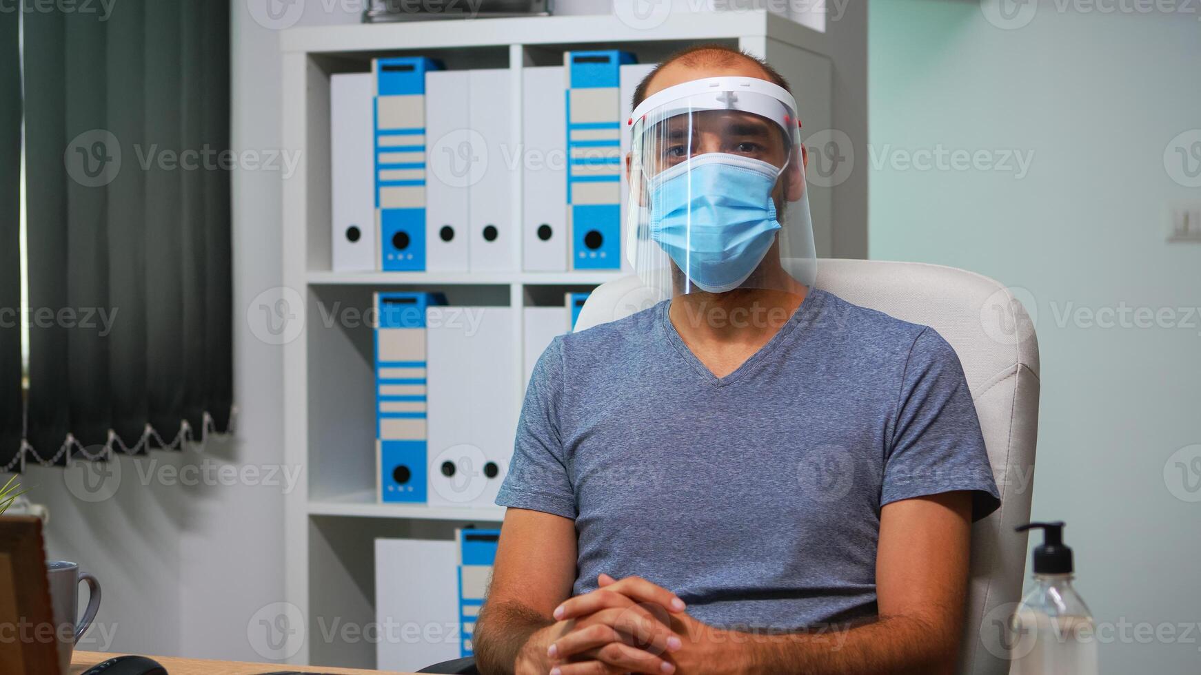 Busy employee wearing mask and visor in work place, typing and looking at camera. Entrepreneur in new normal personal corporate office writing on computer keyboard analyzing graphics photo