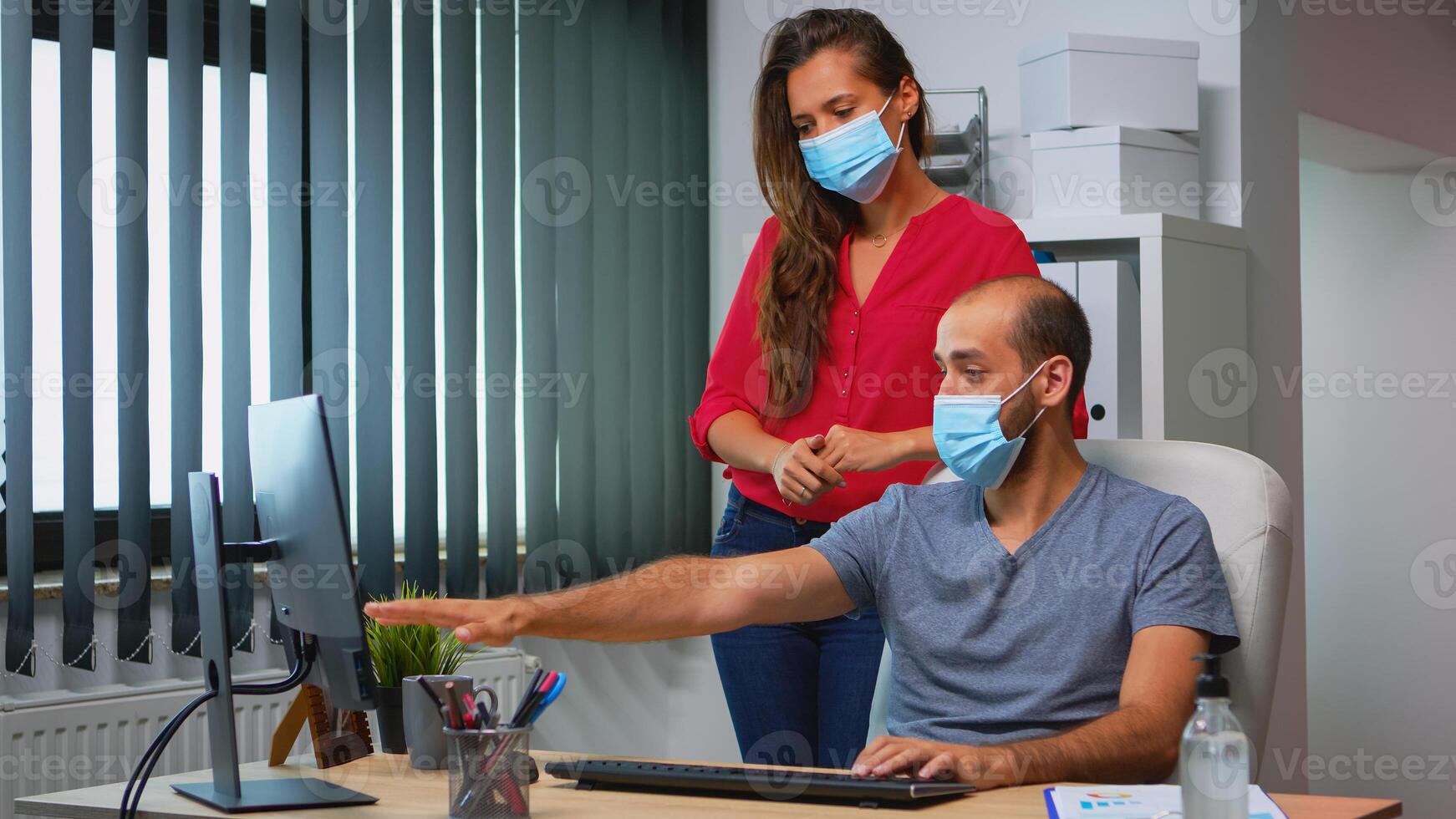 Coworkers with protection face masks working together in workplace during pandemic. Team in new normal office workspace in personal corporate company typing on computer keyboard looking at desktop photo