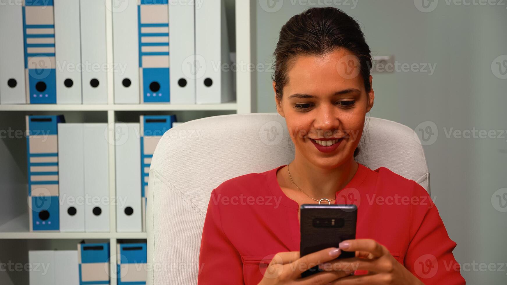 Woman browsing on smartphone smiling sitting on desk in modern office room. Hispanic entrepreneur working in professional workplace in corporate company typing on mobile phone in front of computer photo