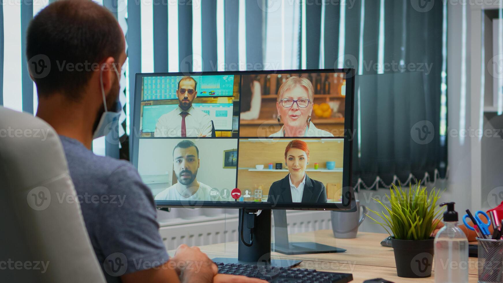 Back view of man with mask talking on video call with remotely colleagues. Freelancer working in new normal office workplace chatting during virtual conference, meeting, using internet technology photo