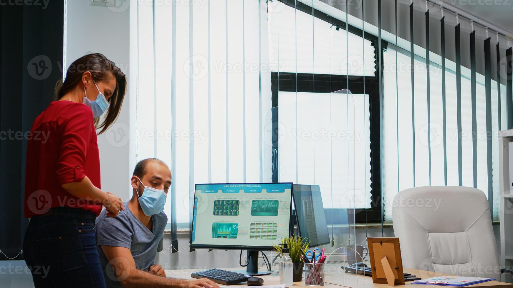 Colleagues with masks discussing graphics sitting in new normal office room. Coworkers talking working pointing at desktop respecting social distancing against covid virus using plexiglass. photo