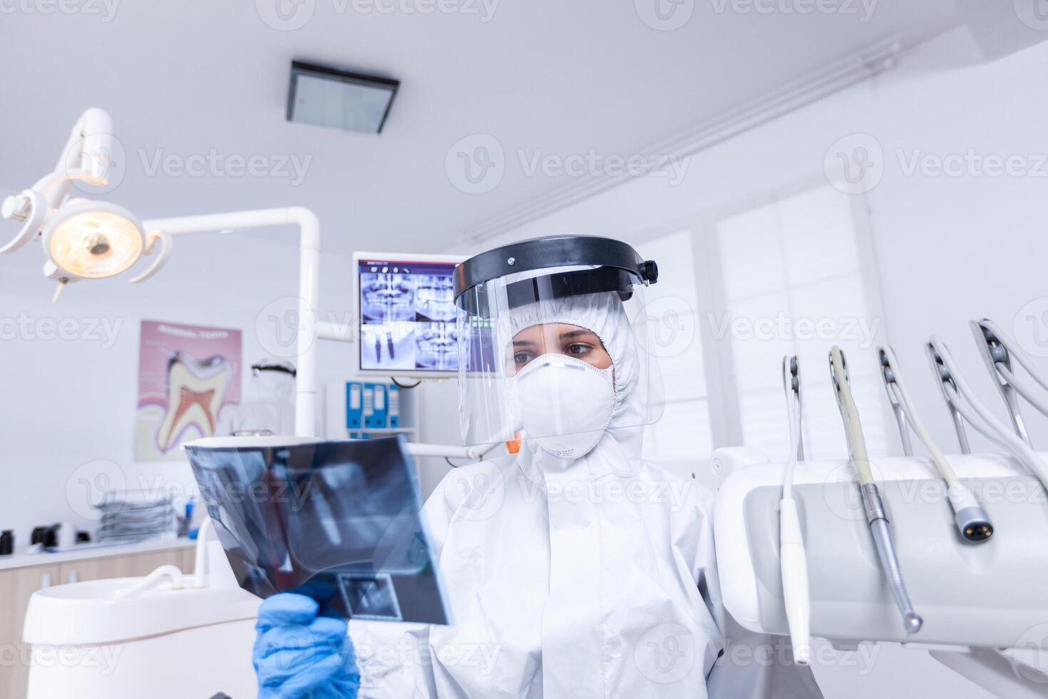 Patient pov in dental office looking at dentist holding radiography dressed in protective gear during covid-19. Dental specialist wearing protective hazmat suit against coroanvirus showing radiography. photo