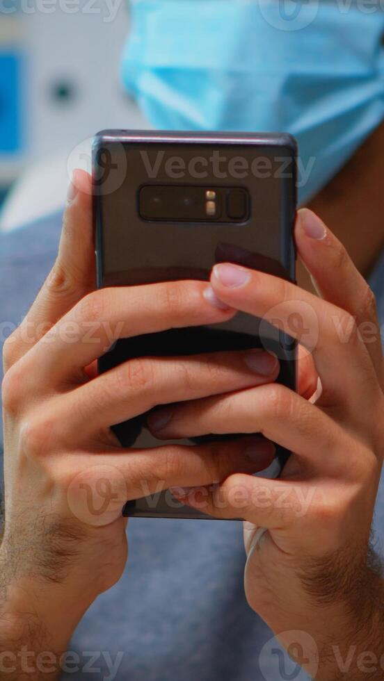 Close up of man with face mask typing text messages on smartphone sitting in workplace during coronavirus. Freelancer working in new normal office chatting talking writing using internet technology photo