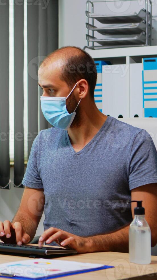 Employee with mask cleaning hands with antibacterian gel in office room before working at computer. Entrepreneur working in new normal office workplace using sanitize alcohol gel against corona virus photo