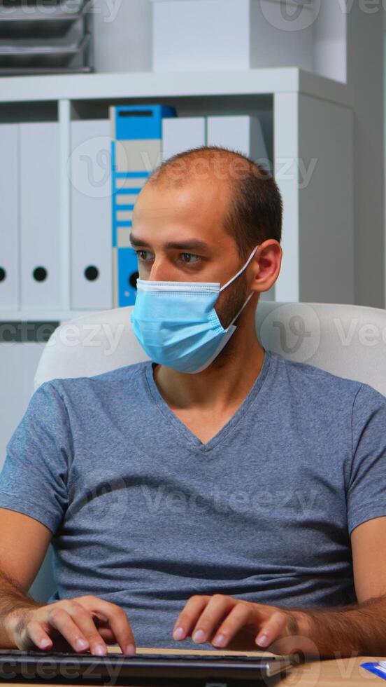 Young manager with protection face mask working alone in office during social distancing. Entrepreneur in new normal personal workplace corporate writing on computer keyboard looking at desktop photo