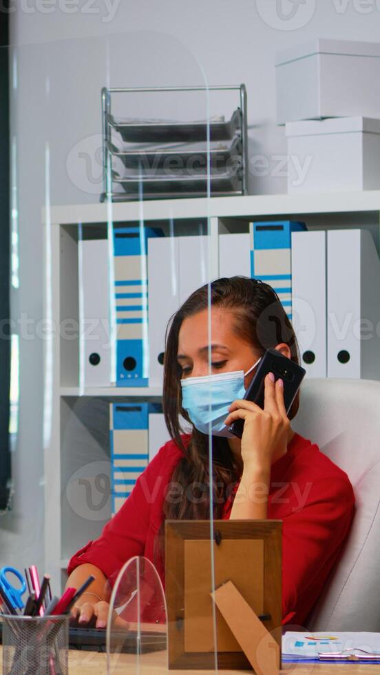 Woman with face mask talking on phone looking at desktop and analyzing the statistics. Freelancer working in workplace chatting with remotely team speaking on smartphone in front of computer photo