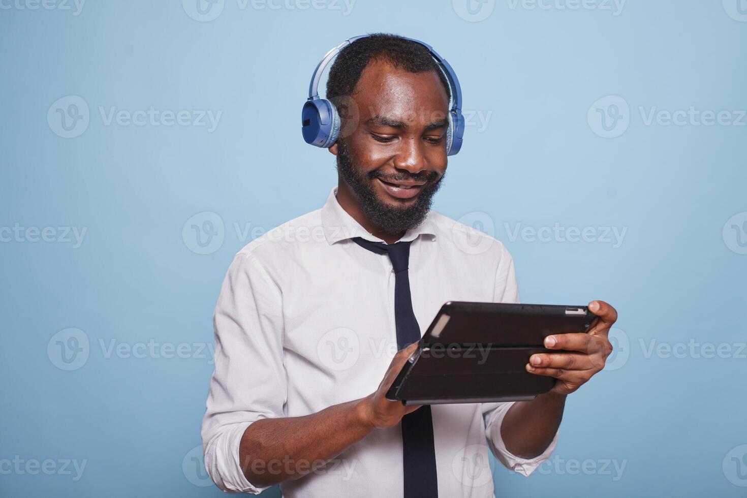 Male entrepreneur relaxing with wireless headphones looking at online show on table against blue background. African american student browsing social media on touchscreen digital device. photo
