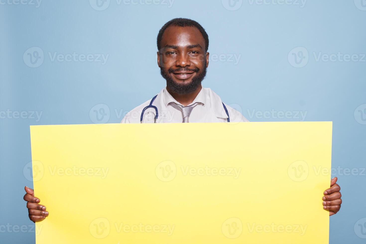 Smiling black doctor with stethoscope grasping large yellow banner for hospital advertising concept. Medical specialist presenting panel with empty copy space over isolated blue background. photo