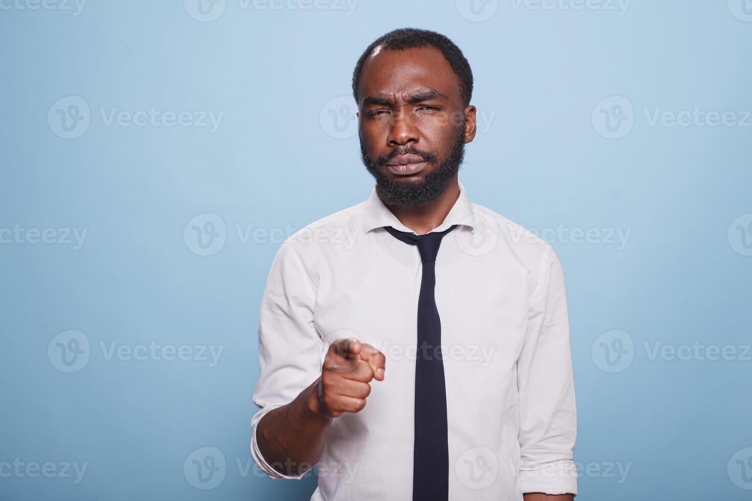 Businessman is standing and looking puzzled while pointing to his front, trying to recognize a person. Confused office worker narrowing his eyes and gestures with his index finger towards camera. photo