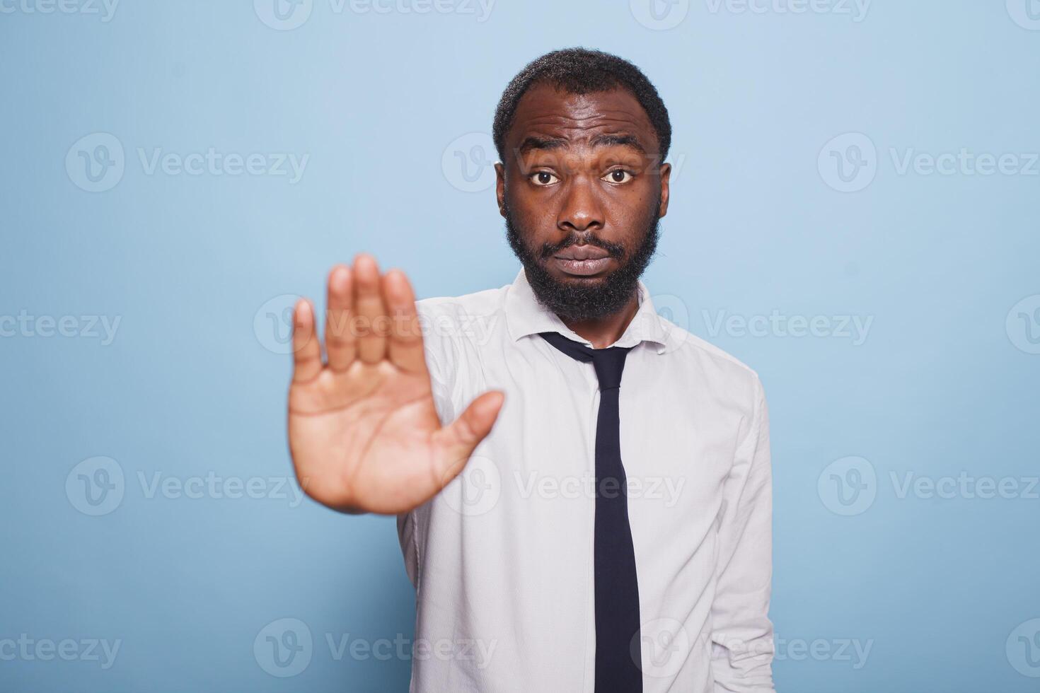On isolated blue background, serious looking businessman in a white shirt and black tie, makes a hand stop in disapproval. Professional office worker with a grumpy demeanor putting up denial sign. photo