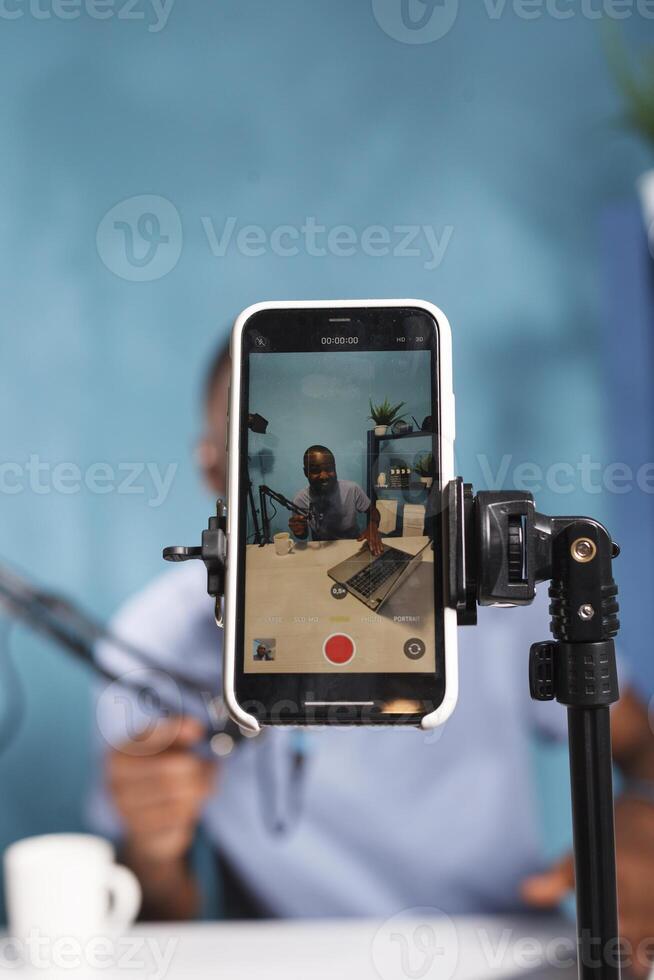 Blogger on smartphone screen using laptop and recording video closeup. Smiling african american web content creator live streaming with mobile phone and looking at camera photo