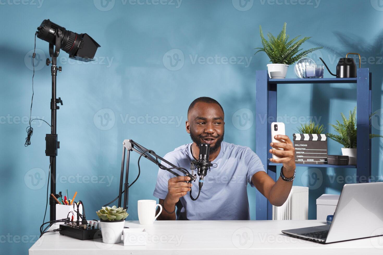 African american vlogger holding smartphone and speaking in microphone while creating web content. Young smiling blogger streaming on social media channel using mobile phone photo