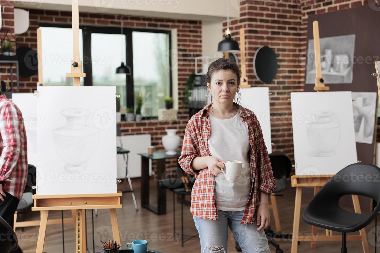 People and creative hobbies. Young woman standing against wooden easels in art classroom holding cup of coffee or tea, people learning pencil sketching techniques at school of contemporary arts photo
