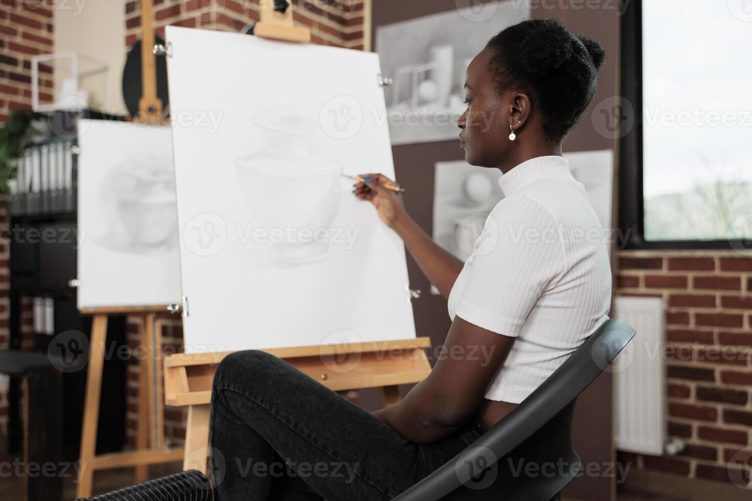 Young focused African American woman learning to draw at group drawing class, sitting at easel in classroom and sketching object with pencil on canvas, people taking up art classes for stress relief photo