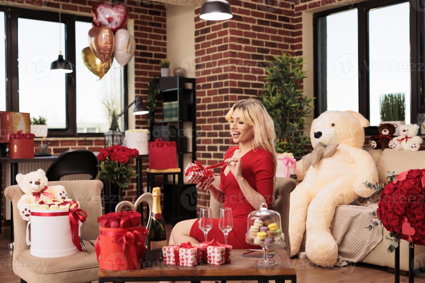 Cute girlfriend sitting in living room filled with romantic gift, enjoying celebrating love holiday. Attractive sexy woman in red dress receiving valentine s day presents from boyfriend photo