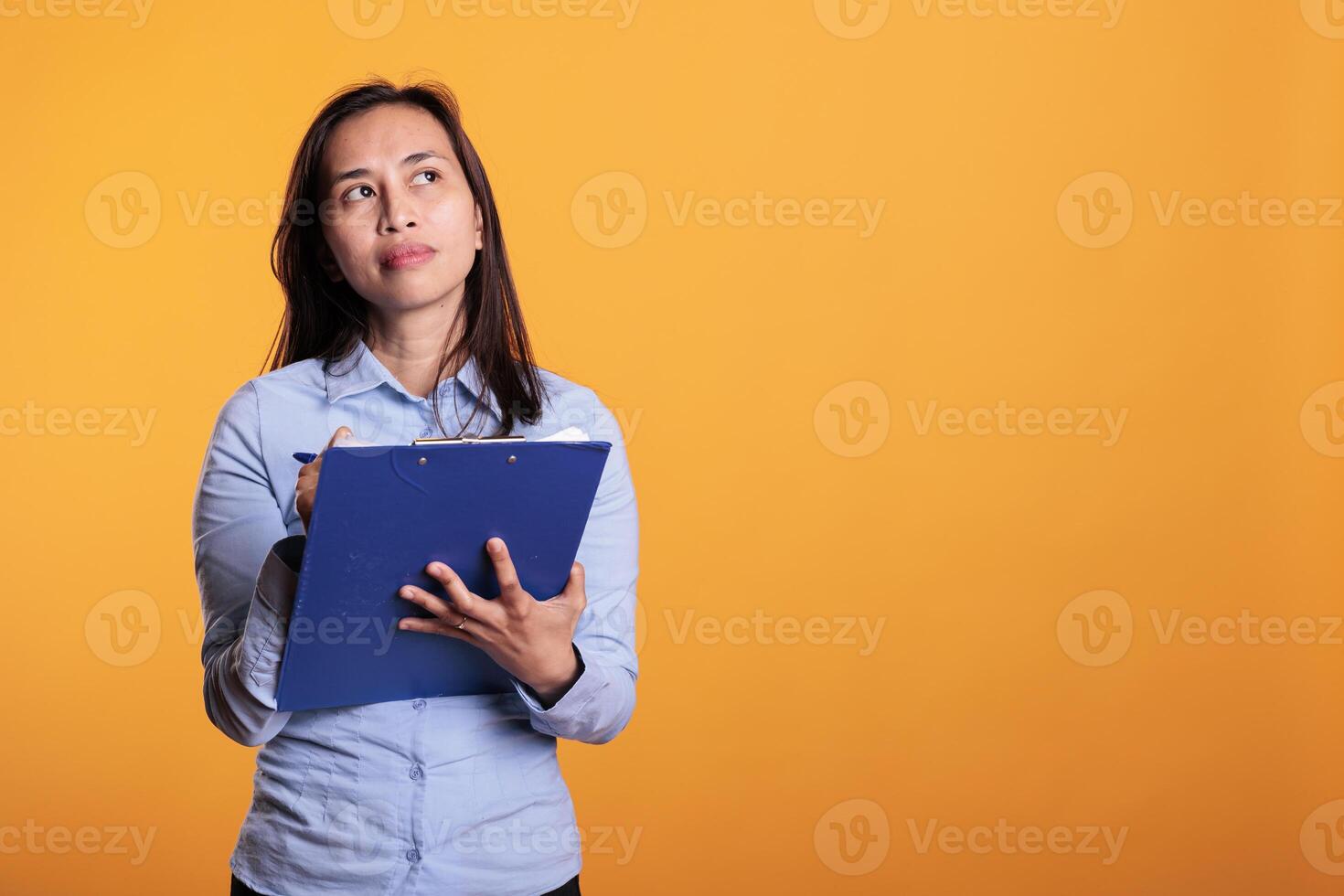 Filipino woman brainstorming ideas for business project, writing report on documents. Pensive thoughtful model looking at checklist, answering questionnaire in studio over yellow background photo