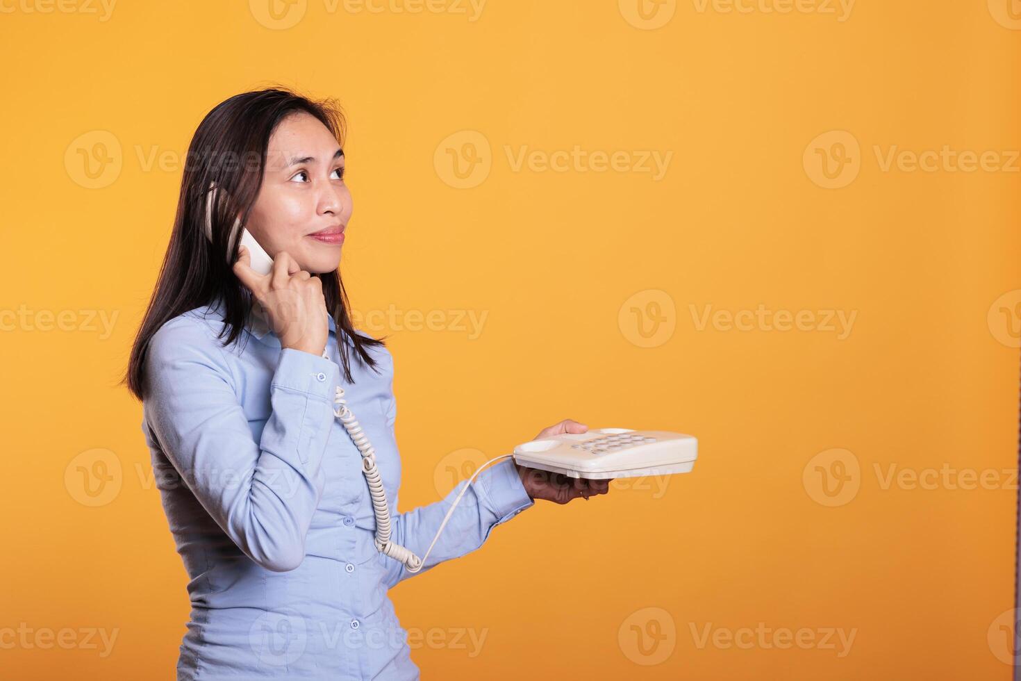 positivo asiático mujer teniendo remoto conversacion con amigo, hablando a teléfono fijo teléfono en estudio terminado amarillo antecedentes. grave adulto respondiendo llamada que se discute acerca de negocio proyecto con compañero de trabajo foto