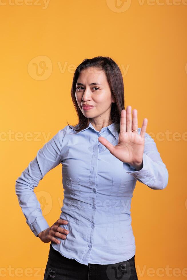 Serious asian woman raising palm to advertise stop gesture in studio over yellow background, expressing denial and refusal. Negative young adult showing rejection gesture, doing rejection symbol photo