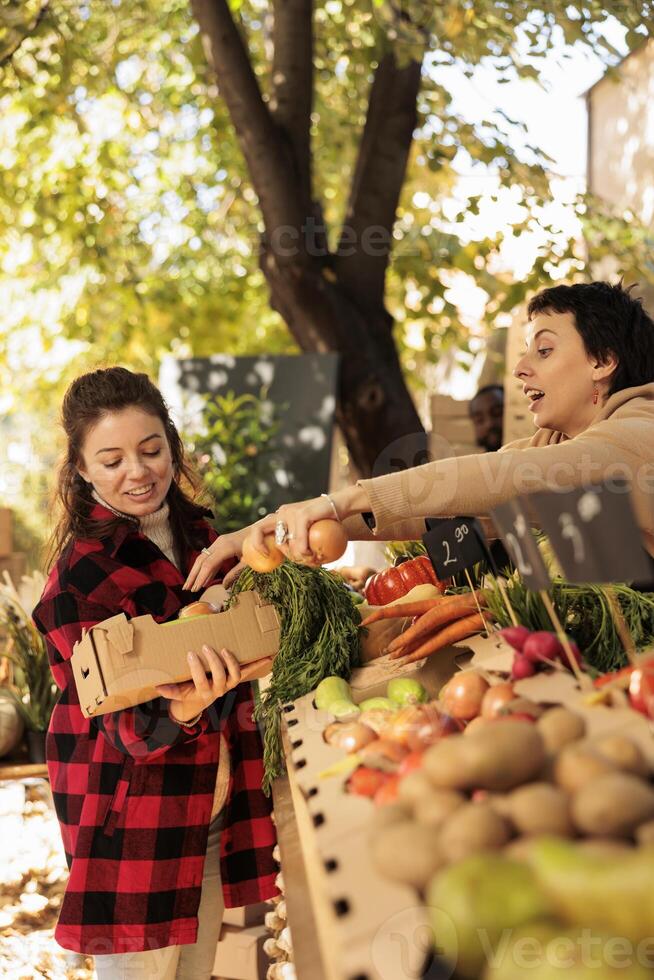 Young happy millennial woman buying fresh organic vegetables at local farmers market, smiling female customer holding box full of seasonal veggies while standing near stall with various farm products photo
